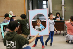 長方形に丸い穴が開いている枠を抱えた大人と、その穴から顔を出している子どもの写真