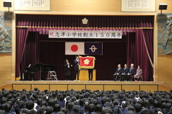 体育館の舞台上で「祝　志津小学校創立150周年」の看板と校旗が掲げられているようす