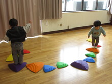 室内で、床の上のバランスストーンに乗りバランスをとるこどもたちの写真